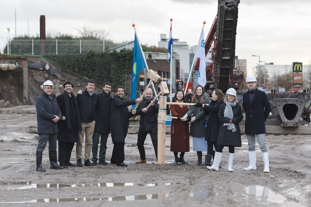 Een groep mensen staat op een bouwplaats, rondom een houten paal die symbolisch de grond in wordt geslagen bij de start van de bouw van project Kop Dakpark