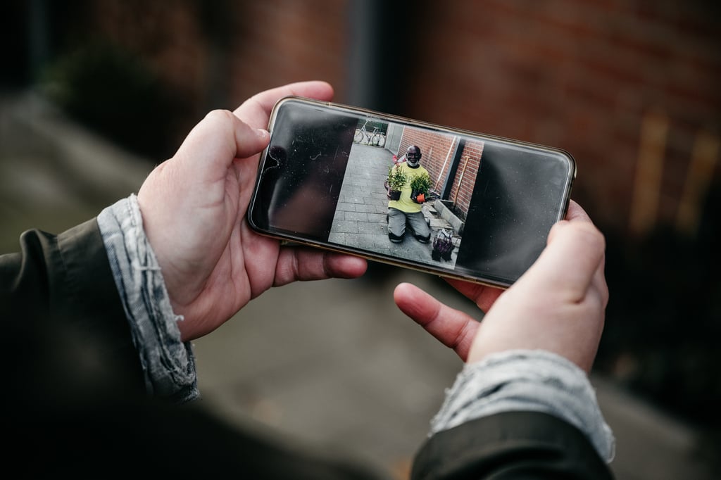 Iemand houdt een smartphone vast, waarop we het beeld zien van iemand geknield zit met twee planten in zijn handen.