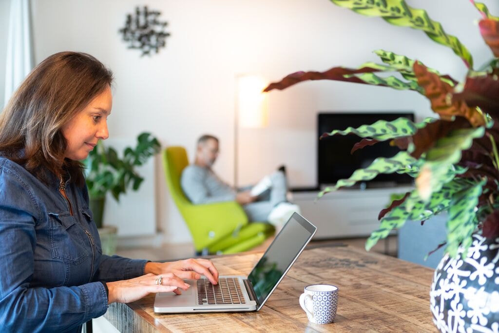 Bewoner zit aan tafel met een laptop voor haar. Op de achtergrond, in de woonkamer, zien we een man in een stoel zitten.