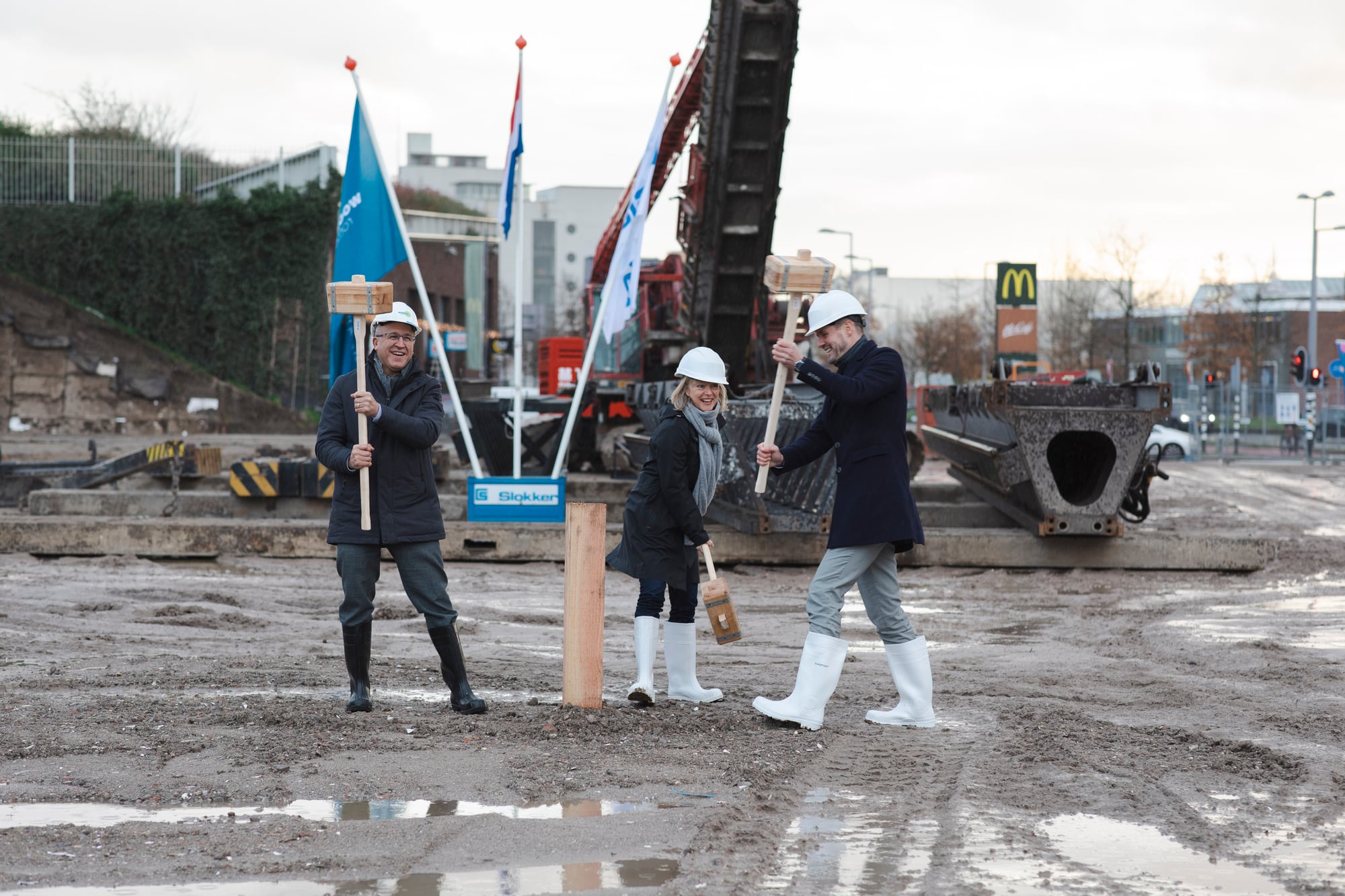 Drie mensen staan op een bouwplaats, en slaan met grote hamers een houten paal die symbolisch de grond in wordt geslagen bij de start van de bouw van project Kop Dakpark